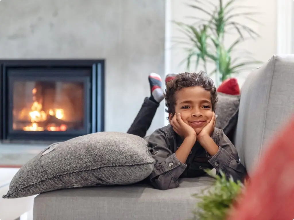 Boy lying on stomach on sofa with head in hands, smiling and looking away with gas fireplace in background.