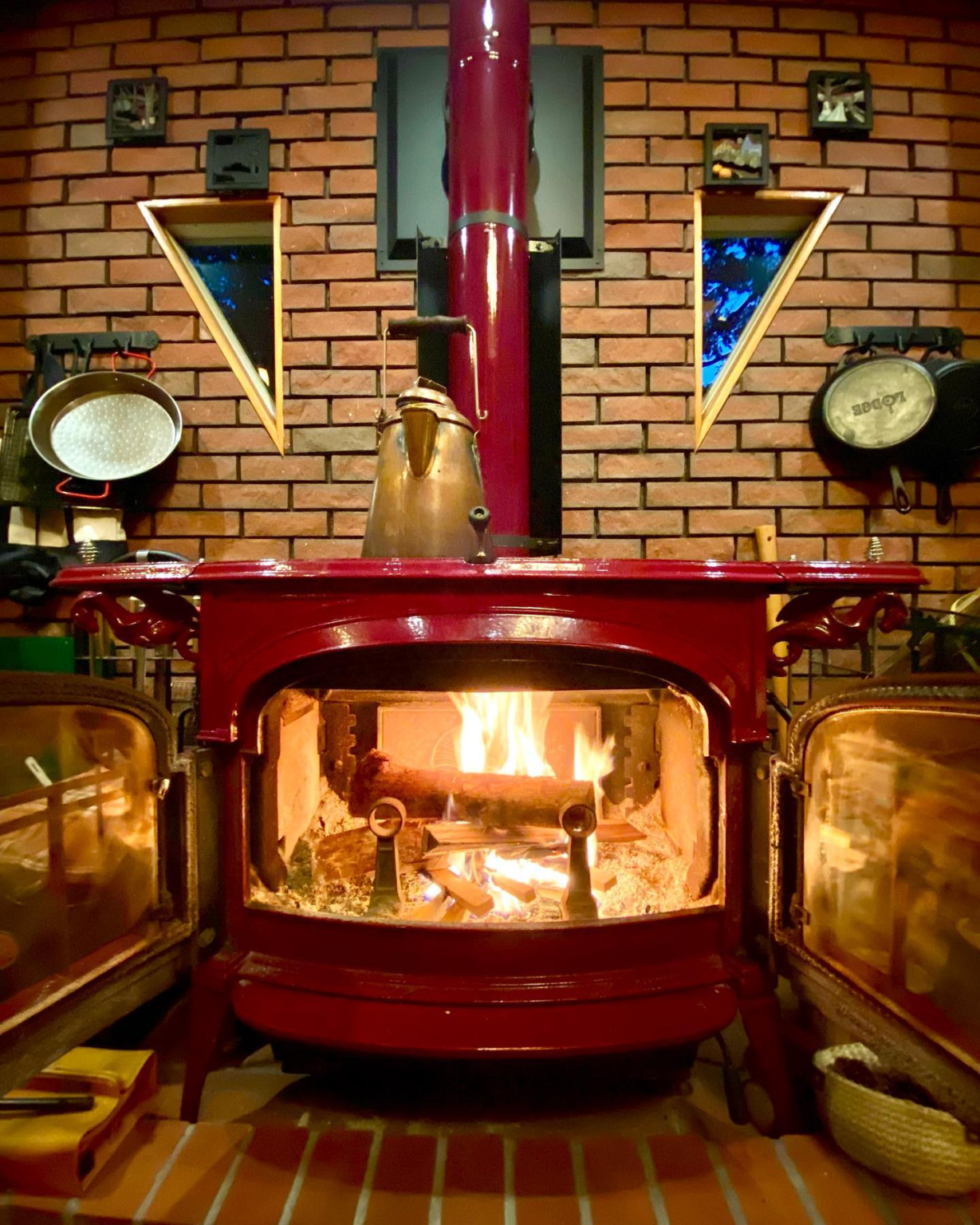 open wood stove vermont castings encore in red with brick wall background closeup of fire wamr heat coming out into living room with a kettle on top and pans hanging on the walls
